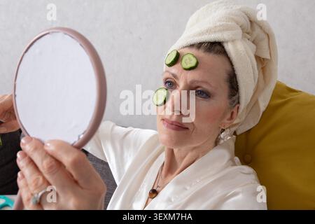 La donna anziana si siede comodamente sul divano avvolto in un asciugamano, applicando la cura della pelle con l'aiuto di fette di cetriolo, appoggiandosi sulla fronte, l Foto Stock