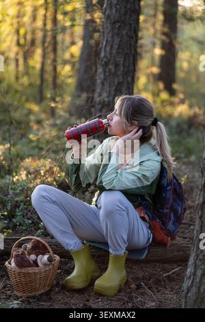 Donna che beve il tè da thermos dopo aver forgiato i funghi nella foresta seduta a riposare. Interruzione idratazione. Foto Stock