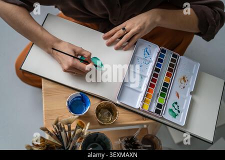 Artista donna che dipinge con acquerelli in un libro di schizzi a casa, tecnica di apprendimento e media. Foto Stock