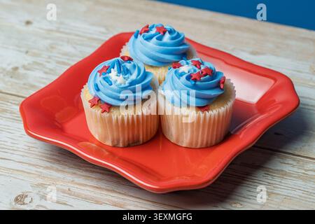 Tre cupcake con glassa blu e stelle rosse si trovano su un piatto rosso. Tavolo in legno e sfondo blu completano la scena. I cupcake sono in carta Foto Stock