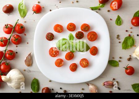 Vista dall'alto dei pomodori ciliegini a fette con basilico fresco su un piatto bianco, circondato da aglio, erbe e spezie su un rustico tavolo in legno. Foto Stock