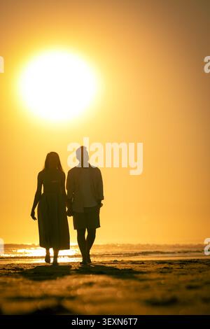 La coppia cammina mano nella mano sulla spiaggia durante il tramonto vicino all'acqua con il sole che tramonta all'orizzonte Foto Stock