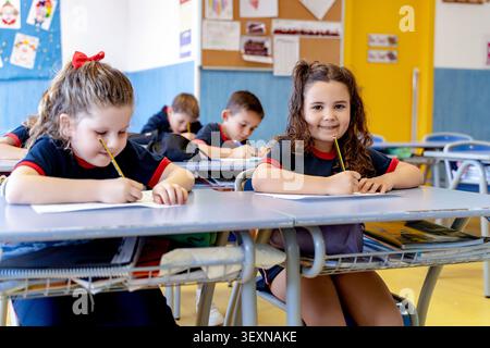 Studenti elementare felici che indossano l'uniforme, si siedono alla scrivania, scrivono su carta e studiano durante la lezione scolastica Foto Stock