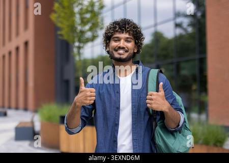 Giovane indiano felice con zaino e capelli ricci che danno un gesto con i pollici, esprimendo il successo e le emozioni positive all'aperto nel campus universitario Foto Stock