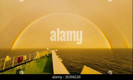 Ampio colpo orizzontale da un ponte di una spettacolare e luminosa arcobaleno doppio che forma un arco perfetto sopra l'oceano aperto scuro sotto una spettacolare S dorata Foto Stock