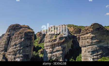 Colpo di droni orizzontale di un antico monastero ortodosso in piedi su un'alta scogliera tra le massicce formazioni rocciose di Meteora sotto un cielo limpido e blu Foto Stock