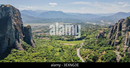 Vista panoramica orizzontale aerea di una splendida valle verde con una strada tortuosa tra le massicce formazioni rocciose di Meteora sotto una montagna lontana Foto Stock