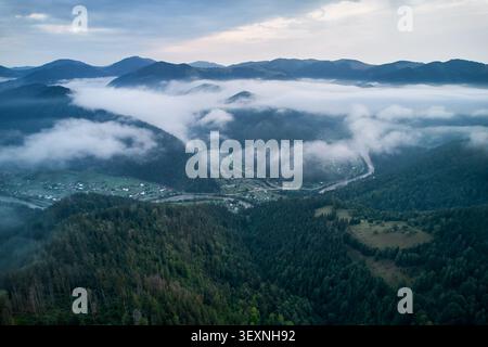 Vista aerea mozzafiato della valle avvolta dalla nebbia mattutina, con il fiume che si snoda tra lussureggianti colline verdi e il villaggio annidato tra gli alberi. Foto Stock