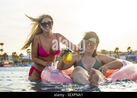 Due giovani donne che si rilassano in mare al tramonto, una in piedi e sorridente sorridente mentre l'altra galleggia in un gonfiabile Foto Stock
