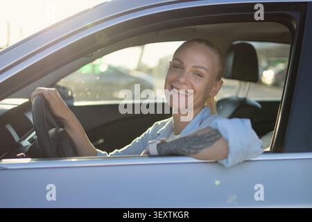 Donna che sorride con sicurezza con un tatuaggio sul braccio, guida un'auto in una giornata di sole, rappresenta lo stile di vita moderno, l'indipendenza e la gioia della persona Foto Stock