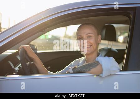 Una giovane donna sorridente mentre è seduta in macchina, tenendo il volante con una mano e appoggiando il braccio tatuato sulla finestra aperta, es Foto Stock