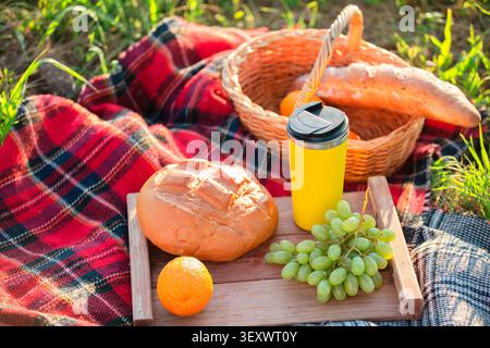 Una giornata di sole si svolge mentre un picnic mette in mostra pane fresco, uva e un'arancia, accompagnati da una bevanda gialla, il tutto riposato su una coperta accogliente Foto Stock