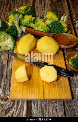 Le patate pelate e i broccoli freschi sono pronti per essere preparati su un tagliere di legno con un coltello e cucchiai di legno. Foto Stock
