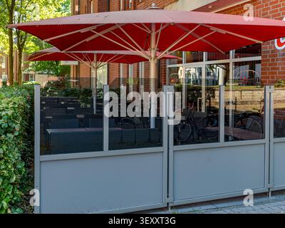 Terrazza all'aperto vuota di un caffè con grandi ombrelloni rossi, pannelli frangivento in vetro e sedie intrecciate accanto a un edificio in mattoni Foto Stock