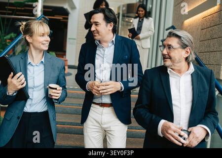 Donna d'affari sorridente che parla con colleghi mentre si sposta giù per le scale nell'edificio degli uffici Foto Stock