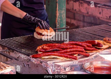 Uomo che prepara un hamburger in un mercato di campagna e grigia salsicce rosse brillanti su una griglia piatta Foto Stock