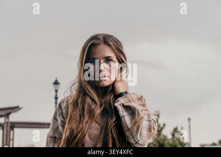 Occhiali da donna ritratti giovane donna dai capelli lunghi che indossa una camicia a quadri all'aperto contro un cielo neutro Foto Stock