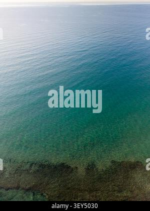 La prospettiva con droni ad alto angolo cattura l'acqua cristallina dell'oceano turchese con piccole increspature che rivelano una struttura rocciosa scura vicino al fondo. Foto Stock