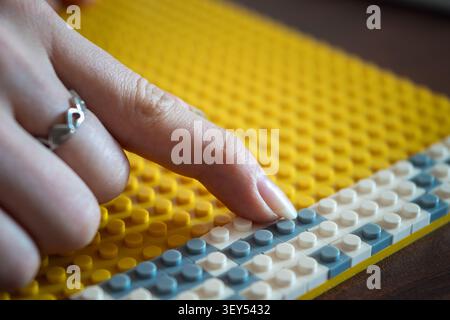 Primo piano di una mano con un anello d'argento che posa una piccola piastrella mosaico bianca su una tavola a pioli gialla in plastica. Arte mosaico creativa, motricità fine ed educazione Foto Stock
