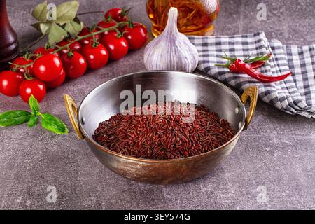 Riso rosso in una ciotola di metallo vintage circondata da pomodori ciliegini aglio peperoncino olio d'oliva basilico su una superficie grigia testurizzata. Luci calde r Foto Stock