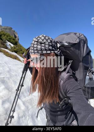 Escursionista femminile con zaino e bastoni da trekking cammina lungo il sentiero innevato. Giornata di sole con cielo azzurro, paesaggio alpino primaverile durante il periodo chal Foto Stock