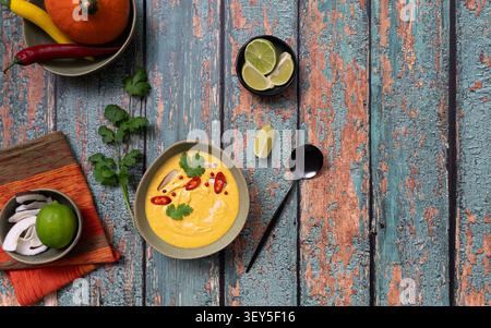 Zuppa di zucca colorata servita con peperoncino, spicchi di lime ed erbe fresche su un rustico tavolo di legno Foto Stock