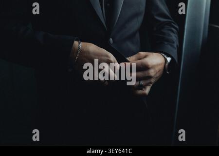 Vestito con un abbigliamento formale scuro, l'uomo adatta il suo vestito. Le sue mani mostrano un'attenta attenzione ai dettagli e alla precisione. L'illuminazione mette in risalto i suoi accessori Foto Stock