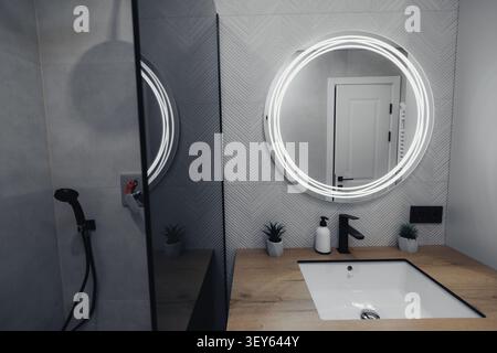 Bagno moderno con specchio LED circolare e lavandino minimalista. Piastrelle grigie e ripiani in legno creano un'atmosfera serena e contemporanea. Finiture nere e piccole Foto Stock