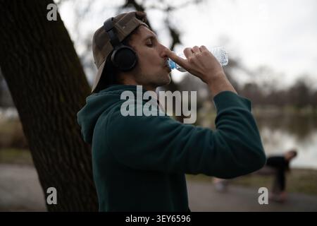 Il giovane beve acqua mentre indossa le cuffie e un berretto in un parco Foto Stock