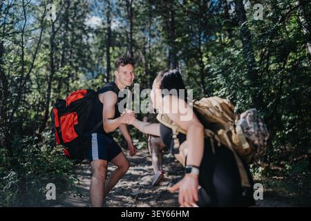 Giovani escursionisti che si aiutano a vicenda lungo un sentiero nella foresta con zaini e sorrisi Foto Stock