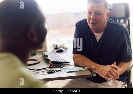 Medico di sesso maschile seduto alla sua scrivania e a parlare con il paziente durante la consultazione Foto Stock