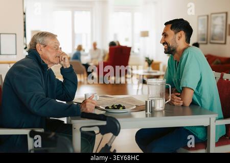 Infermiere maschio che parla con un uomo anziano seduto a tavola nella casa di riposo Foto Stock