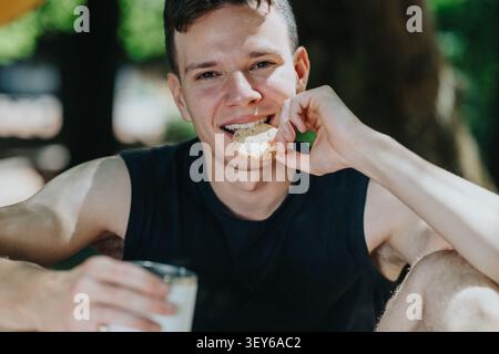 Un giovane che fa escursioni all'aperto sorseggiando un biscotto e un bicchiere di latte mentre sorride e riposa Foto Stock