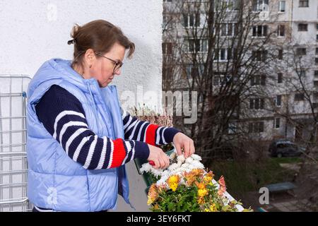 Una donna di 50 anni tende a fiori autunnali sul suo balcone. Si concentra su ogni pianta con cura mentre è circondata da edifici cittadini. È un pomeriggio fresco nella stagione autunnale Foto Stock
