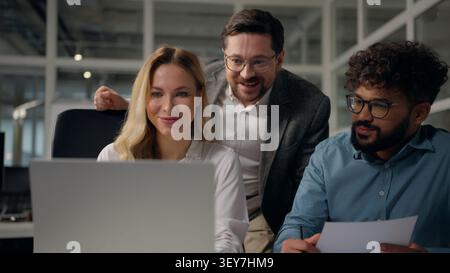 Tre colleghi di uffici aziendali diversificati sorridenti che guardano in laptop che parlano allegri che godono di buoni risultati online parlano colleghi multietnici Foto Stock