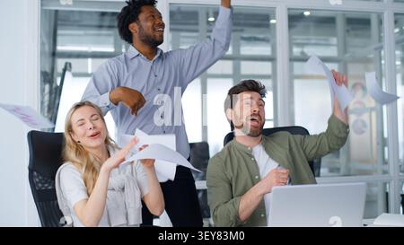 Gruppo aziendale tre colleghi multirazziali che ballano lanciano documenti che celebrano il fine settimana di lavoro completo cantando felici nella diversità degli uffici Foto Stock