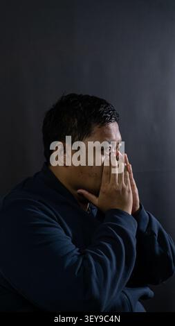 Un profilo laterale ravvicinato di un maschio indonesiano ansioso che stringe entrambe le mani davanti alla bocca mostra profonda preoccupazione, lotta interna e alto st Foto Stock