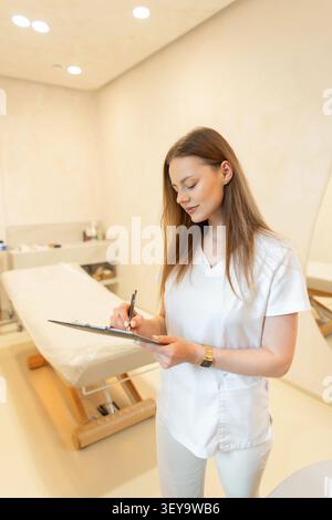 Giovane cosmetologa donna o specialista medico in scrub bianchi compilando la scheda paziente su un clipboard, in piedi in una luminosa clinica di benessere moderna Foto Stock