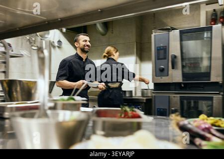 Uno chef sorridente che cammina in cucina mentre tiene il piatto in cucina Foto Stock
