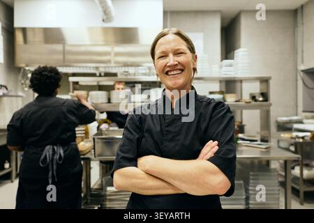 Ritratto di una allegra chef donna in piedi con le braccia incrociate nella cucina del ristorante Foto Stock