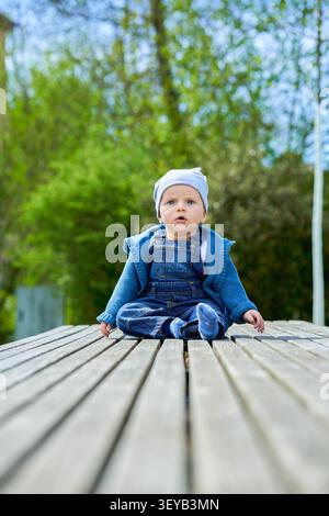 Ragazzo fino a un anno di età, aspetto europeo, occhi blu si siede in estate su una panchina di legno vestita con tute in denim e un berretto leggero. Alta qualità Foto Stock