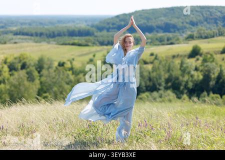Una donna serena in un abito blu che scorre medita all'aperto, abbracciando la tranquillità della natura e promuovendo il relax e la consapevolezza in una lussureggiante lan verde Foto Stock