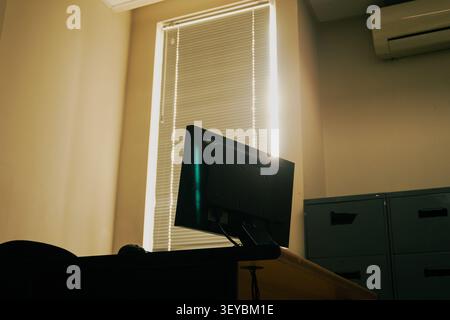Il monitor del computer è rivolto verso una sedia vuota senza presenza di persone. Le tende verticali filtrano la luce del giorno dal vento alto Foto Stock