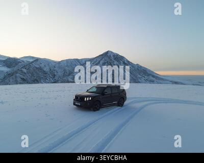 Bishkek, Kirghizistan - 18 dicembre 2022: Vista laterale ad angolo alto di un SUV di lusso nero Kia Mohave che si muove lungo una pista innevata con una maestosa vetta di montagna Foto Stock