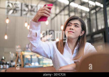 Ritratto di uno stile di vita giocoso di una donna transgender isana del sud-est asiatico, che ha compiuto 40 anni, scattando selfie e guardando in alto con il suo telefono in un bar restaura Foto Stock