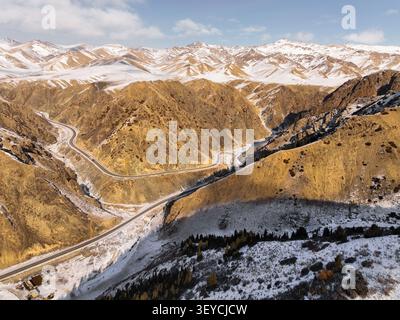 Ampio panorama aereo di una tortuosa strada lastricata che attraversa un aspro e arido fondovalle con creste di montagna innevate e colline ai piedi del distan Foto Stock