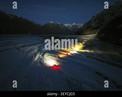 Vista aerea ad angolo basso che mostra le luci dei veicoli fuoristrada illuminando in modo luminoso il fondo innevato della valle sotto un cielo blu scuro Foto Stock