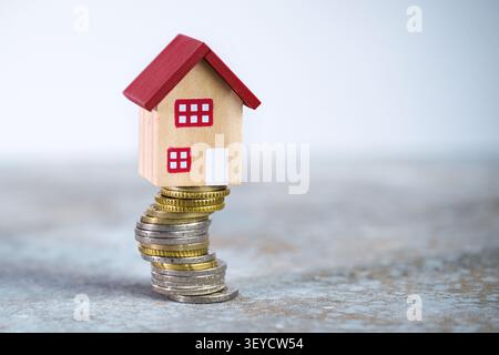 Modello di casa arroccato su un'instabile e alta pila di monete, concetto di fluttuazione dei costi di costruzione, aumento dei tassi di mutuo o diminuzione del valore di un pr Foto Stock