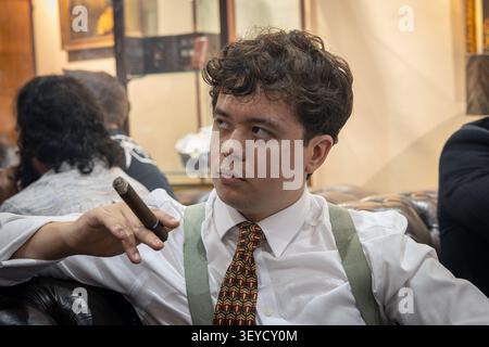 Ritratto di un giovane uomo sicuro di sé che indossa l'apparecchio sopra una camicia bianca che fuma un sigaro di lusso. Foto Stock