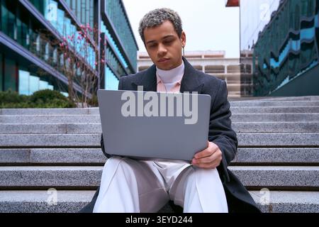 Il bel giovane afroamericano vestito con un abbigliamento alla moda lavora sul suo laptop all'aperto. Foto Stock
